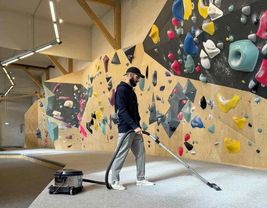 Person vacuuming climbing mats beneath an indoor bouldering wall.